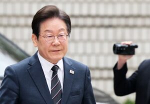 Middle‑aged man in a dark suit and striped tie, wearing a small flag pin, with a handheld video camera visible in the background.